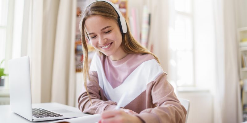 Young happy college female student sitting at the table at home, using laptop and headphones when studying.